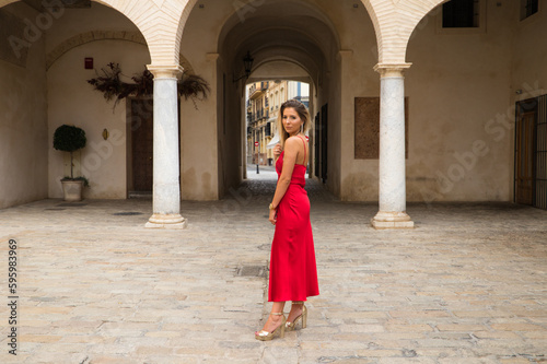 Young, blonde, beautiful woman in a red dress is visiting seville. The woman poses for the camera very elegant and like a model in the typical streets of the city. Holidays and travels © @skuder_photographer