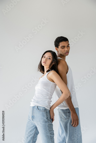 brunette asian woman in jeans and tank top looking at camera while standing back to back with african american man isolated on grey.