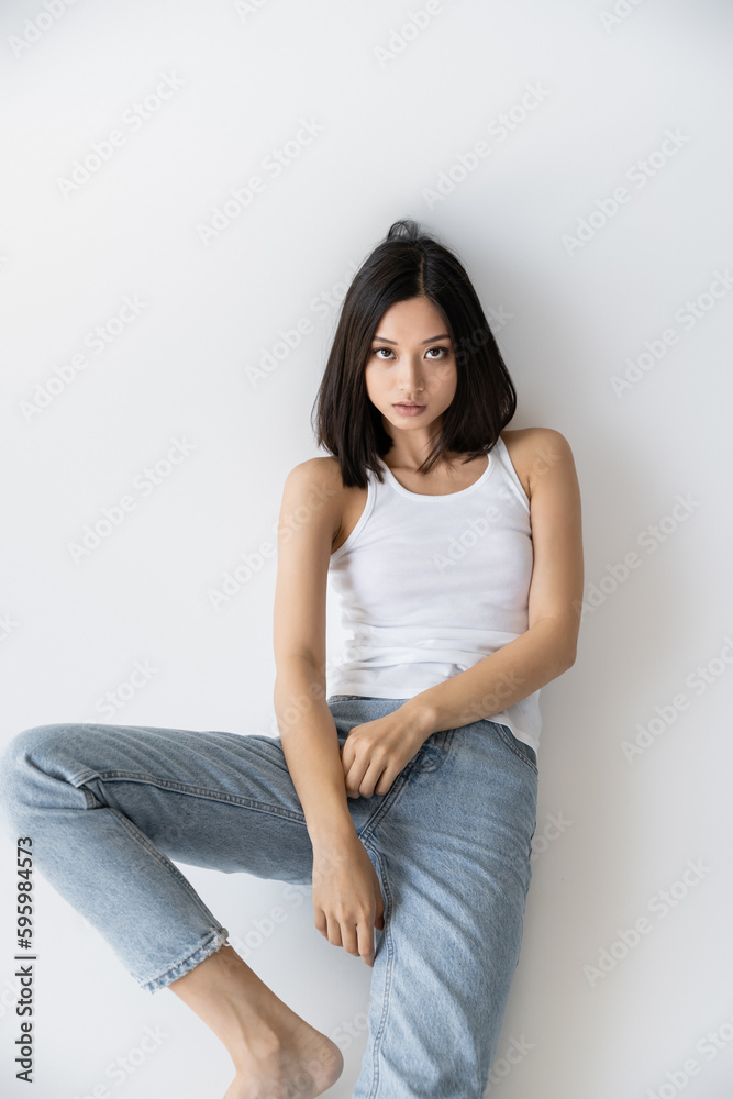 brunette asian woman in blue jeans and white tank top sitting and looking at camera on grey background. brunette asian woman in blue jeans and white tank top sitting and looking at camera on grey background.