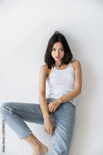 brunette asian woman in blue jeans and white tank top sitting and looking at camera on grey background.