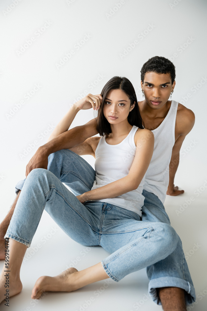 stylish interracial models in blue jeans and white tank tops sitting and looking at camera on grey background. stylish interracial models in blue jeans and white tank tops sitting and looking at camera on grey background.