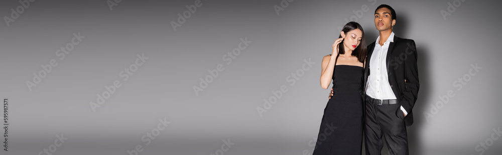 african american guy in black suit standing with hand in pocket near asian woman in strap dress on grey background, banner. african american guy in black suit standing with hand in pocket near asian woman in strap dress on grey background, banner.