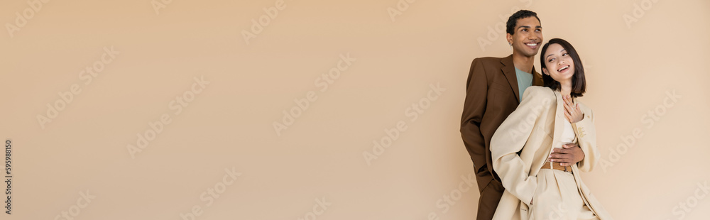 smiling african american man embracing happy asian woman in ivory suit isolated on beige, banner. smiling african american man embracing happy asian woman in ivory suit isolated on beige, banner.