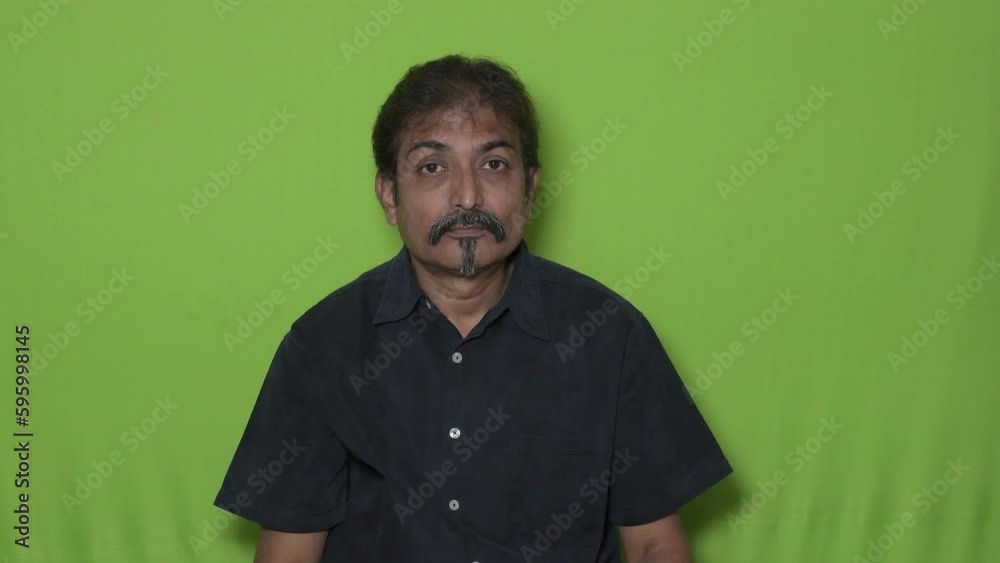 The well-dressed Indian man wearing a black shirt is indicating something to the top direction with his right hand's index fingers while seating against a green screen background The well-dressed Indian man wearing a black shirt is indicating something to the top direction with his right hand's index fingers while seating against a green screen background