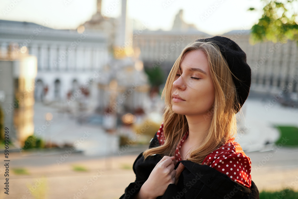 Fototapeta premium Fashion portrait of adorable stylish blonde young caucasian girl in beret and polka dot dress posing outside with urban view at sunset. Elegant concept. High quality image