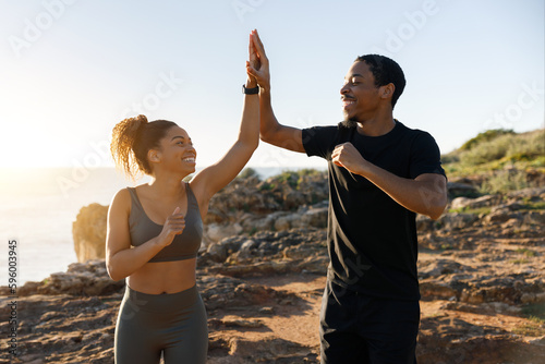 Positive millennial african american husband and wife in sportswear running, give high five on ocean beach