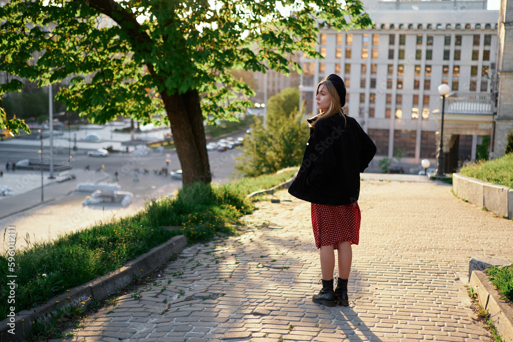 Fashion portrait of stylish elegant lady with urban background at sunset. Attractive blonde young caucasian girl in beret, jacket and polka dot red dress going in the city center. High quality image