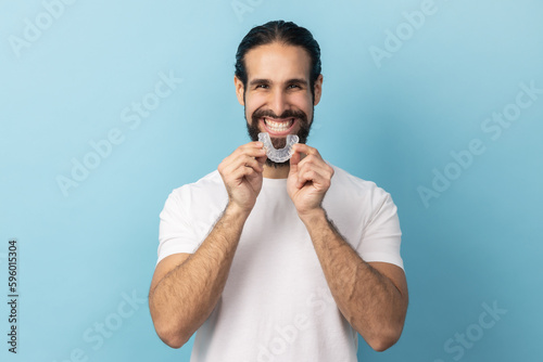 Portrait of man with beard in white T-shirt using teeth whitening braces, dental aligner retainer, dental clinic for beautiful teeth, treatment course. Indoor studio shot isolated on blue background.