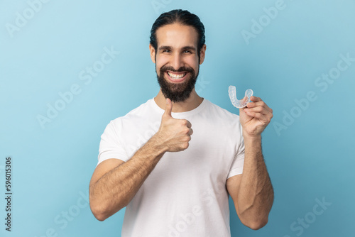 Portrait of man with beard wearing white T-shirt holding dental aligner retainer, dental clinic for beautiful teeth, showing thumb up. Indoor studio shot isolated on blue background. photo