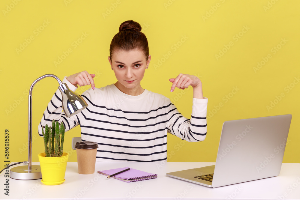 Here and right now. Young woman sitting at workplace with laptop and pointing fingers down, controlling and demanding deadline job finish. Indoor studio studio shot isolated on yellow background. Here and right now. Young woman sitting at workplace with laptop and pointing fingers down, controlling and demanding deadline job finish. Indoor studio studio shot isolated on yellow background.