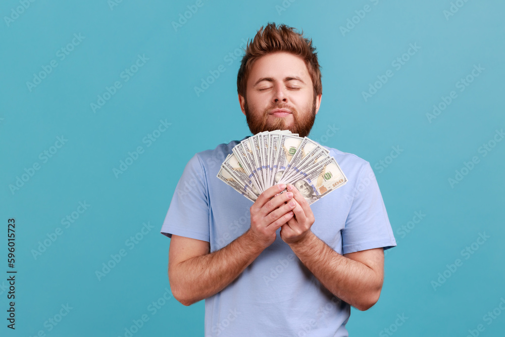 Portrait of satisfied bearded man smelling earned dollar banknotes, enjoying success and big profit, wealthy life, greedy for money. Indoor studio shot isolated on blue background. Portrait of satisfied bearded man smelling earned dollar banknotes, enjoying success and big profit, wealthy life, greedy for money. Indoor studio shot isolated on blue background.
