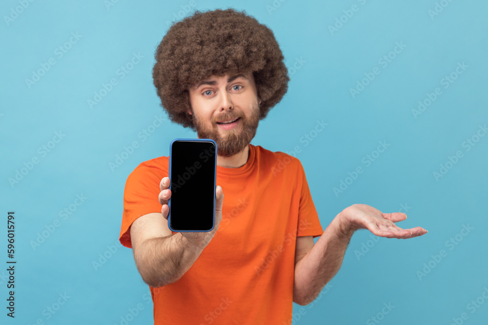 Portrait of confused man with Afro hairstyle wearing orange T-shirt showing cell phone with blank black screen, presenting area for advertisement. Indoor studio shot isolated on blue background. Portrait of confused man with Afro hairstyle wearing orange T-shirt showing cell phone with blank black screen, presenting area for advertisement. Indoor studio shot isolated on blue background.