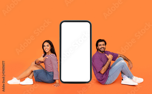 Positive indian couple sitting by big cell phone, mockup