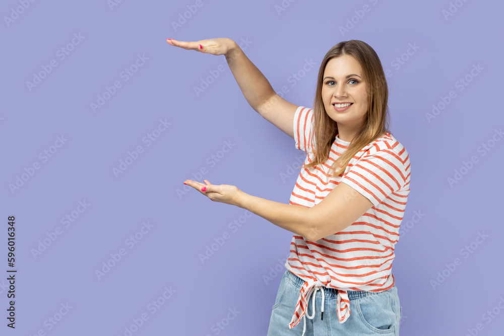 Portrait of satisfied smiling blond woman wearing striped T-shirt presenting area between hands for advertisement, showing huge size. Indoor studio shot isolated on purple background. Portrait of satisfied smiling blond woman wearing striped T-shirt presenting area between hands for advertisement, showing huge size. Indoor studio shot isolated on purple background.