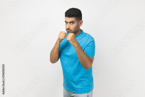 Portrait of confident serious unshaven man wearing blue T- shirt standing with clenched fists, being ready to attack, arguing with somebody. Indoor studio shot isolated on gray background.