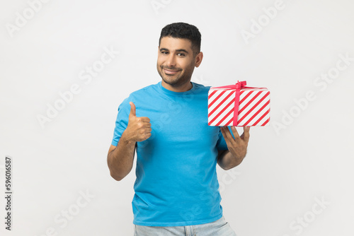 Portrait of attractive joyful unshaven man wearing blue T- shirt standing holding wrapped present box, showing like gesture, thumb up, congratulating. Indoor studio shot isolated on gray background. © khosrork