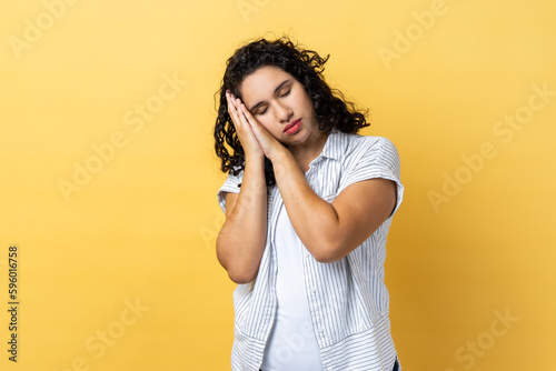 Portrait of exhausted tired beautiful woman with dark wavy hair sleeping laying down on palms  having comfortable nap and resting. Indoor studio shot isolated on yellow background.