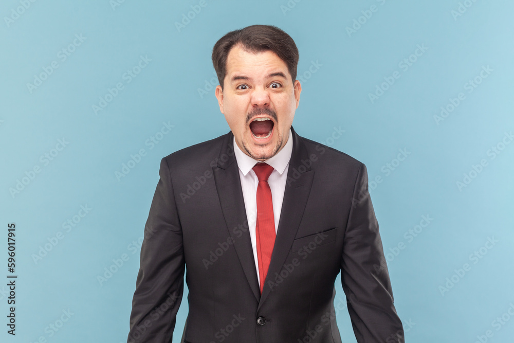 Portrait of angry furious man with mustache standing standing and screaming loud with hate and anger, wearing black suit with red tie. Indoor studio shot isolated on light blue background. Portrait of angry furious man with mustache standing standing and screaming loud with hate and anger, wearing black suit with red tie. Indoor studio shot isolated on light blue background.
