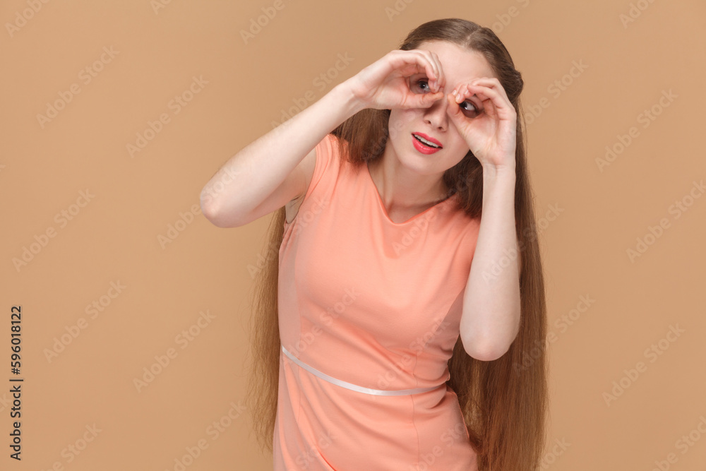 Portrait of funny surprised woman with long hair standing looking at camera through fingers, making binocular gesture, wearing elegant dress. Indoor studio shot isolated on brown background. Portrait of funny surprised woman with long hair standing looking at camera through fingers, making binocular gesture, wearing elegant dress. Indoor studio shot isolated on brown background.