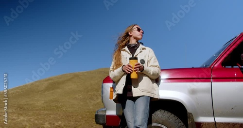 Attractive long-haired girl drinking a drink from a thermos of thermo mugs while standing near a vintage car against the backdrop of a mountain landscape. High quality 4k footage