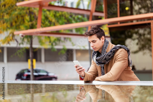 Portrait of stylish handsome young man with coat standing outdoors and leaning on wall using a smartphone
