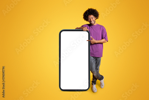 Happy adult african american curly man in purple t-shirt near huge smartphone with blank screen © Prostock-studio
