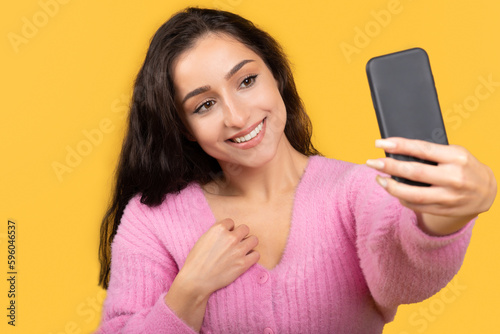 Positive millennial arab woman in casual taking selfie on phone, isolated on yellow background, studio