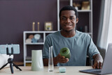 Happy young craftsman with handmade vase in hand looking at camera while sitting in front of laptop and smartphone during livestream