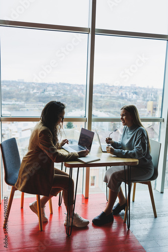 Wallpaper Mural Two young female colleagues sitting with laptops near large windows, modern workspace. Female office workers Torontodigital.ca