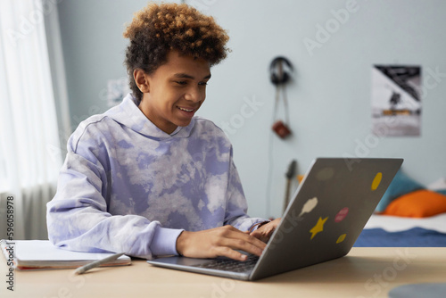 Фотография Smiling diligent teenager in hoodie typing on laptop keyboard while sitting by d