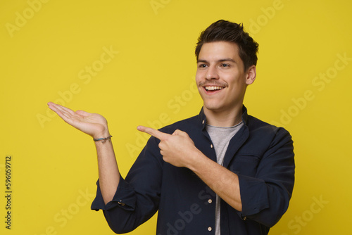 20s young man with excited happy face pointing right by finger to his opened palm and looking to direction isolated over yellow studio background photo