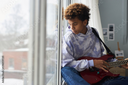 Teenage boy in hoodie and blue jeans playing electric guitar at leisure while sitting on windowsill and looking through window