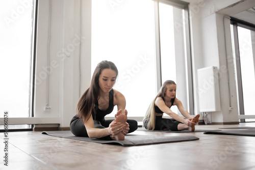Healthy yoga people in sports clothes exercising Janu Sirsasana on sticky mats in fitness studio. Pretty young ladies reducing pain in waist and legs while sitting in Head to Knee Pose during workout.
