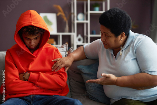 African American mature woman talking to sulky or stressed teenage grandson in red hoodie while sitting on couch next to him photo