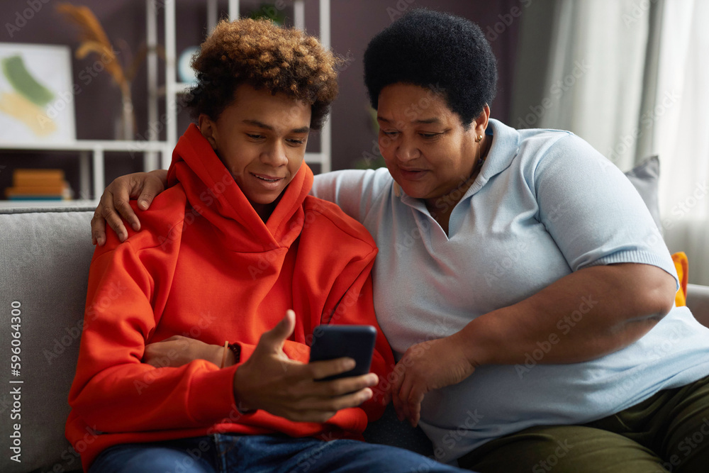 African American teenage boy in red hoodie showing online video in smartphone to his grandmother while both sitting on couch