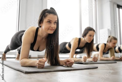 Athletic young females strengthening whole body with Forearm Plank Pose during yoga workout practice in fitness center. Sporty women in activewear improving posture with Phalakasana II exercise.