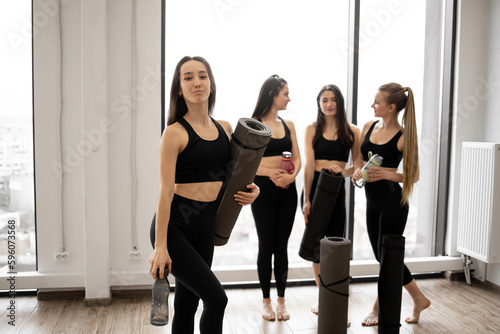 Attractive brunette lady in gym clothes posing with black mat and water bottle while her friends relaxing after workout in sports club. Cheerful female adults promoting health and beauty via yoga.