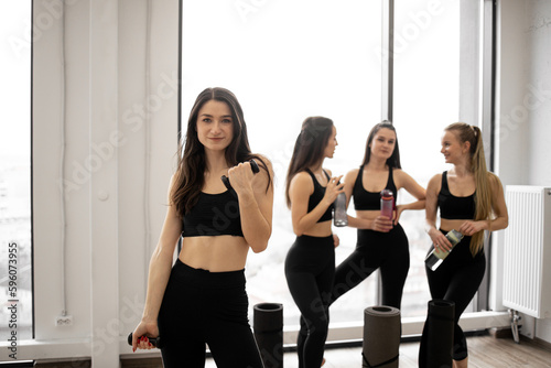 Beautiful slim brunette in activewear lifting dumbbells while fellow yoginis having rest after workout session. Young caucasian ladies with sports equipment talking in fitness studio after exercises.