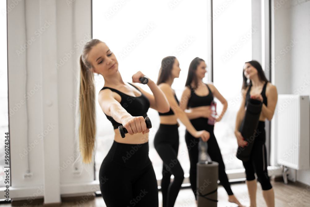 Attractive blonde woman in yoga clothes posing with dumbbells in hands while her female friends relaxing after workout exercises. Cheerful caucasian ladies having good time together in sports club. Attractive blonde woman in yoga clothes posing with dumbbells in hands while her female friends relaxing after workout exercises. Cheerful caucasian ladies having good time together in sports club.