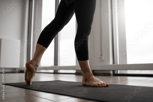Close up view of athletic female in tight pants standing barefoot on black mat while getting into yoga position in bright fitness studio. Sporty woman performing daily workout practice indoors.