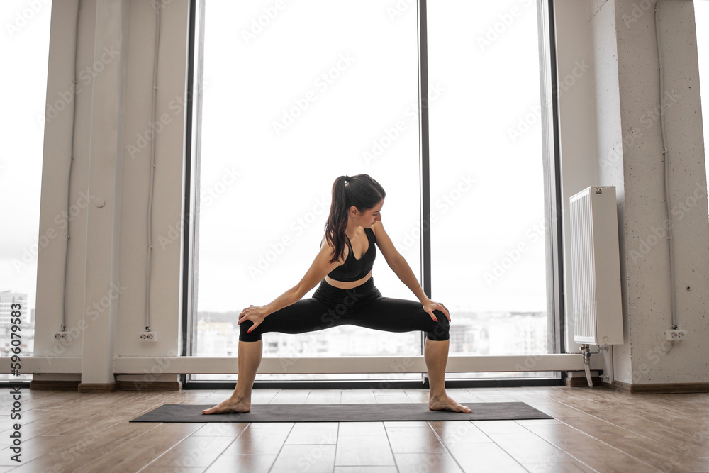 Caucasian woman squatting with feet shoulder width apart, holding hands on hips and twisting torso into sides. Flexible brunette practising sumo squat pose during yoga exercise at studio. Caucasian woman squatting with feet shoulder width apart, holding hands on hips and twisting torso into sides. Flexible brunette practising sumo squat pose during yoga exercise at studio.