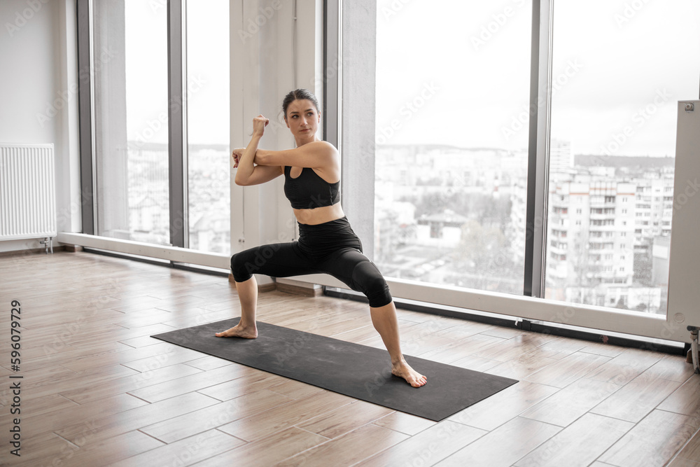 Focused dark haired woman stretching arms and legs during yoga practice at spacious fitness studio. Sporty female wearing black leggings and crop top for training indoors.