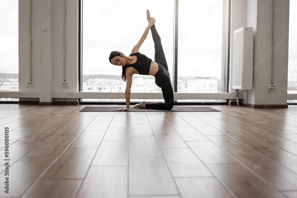 Obraz premium Flexible caucasian woman in black activewear with sportive body doing Ashtanga yoga practice at airy studio with panoramic windows. Concept of spirituality, strenght and meditation.