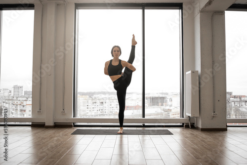 Caucasian dark-haired woman practicing advanced yoga standing in trivikramasana pose in white spacious studio. Professional yogi wearing comfortable sports clothing balancing on one leg doing asana.