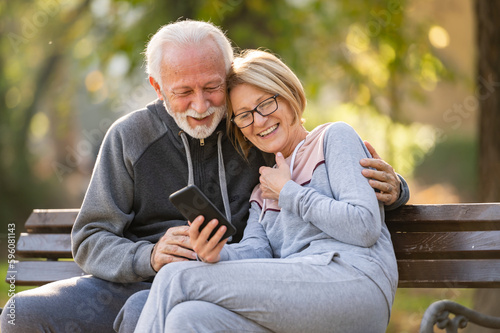 Happy senior couple relaxing together in a city park, sitting on a park bench and using a smart phone