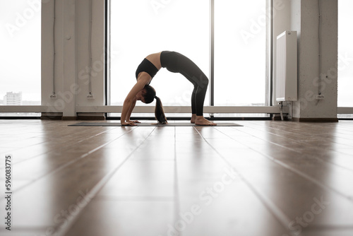 Attractive caucasian yogi dressed in black sport outfit standing in Urdhva Dhanurasana pose during workout. Active woman practicing Bridge exercise on yoga mat at room with french windows.