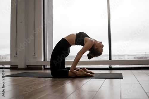 Athletic caucasian woman with bare feet standing in Camel Pose during training at fitness center. Flexible female yogi stretching in Ustrasana exercise with background of French windows.