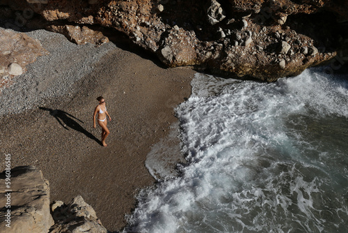 Top view shot of a young woman wearing white bikini swimsuit, alone on a small beach hidden between rocks. Copy space, top view.