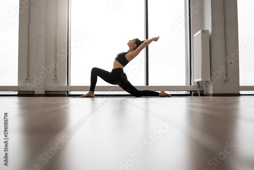 Side view of young sporty female practicing yoga in black outfit at white spacious studio. Slim lady raising hands and arching back near panoramic window. Concept of sport and healthy lifestyle.