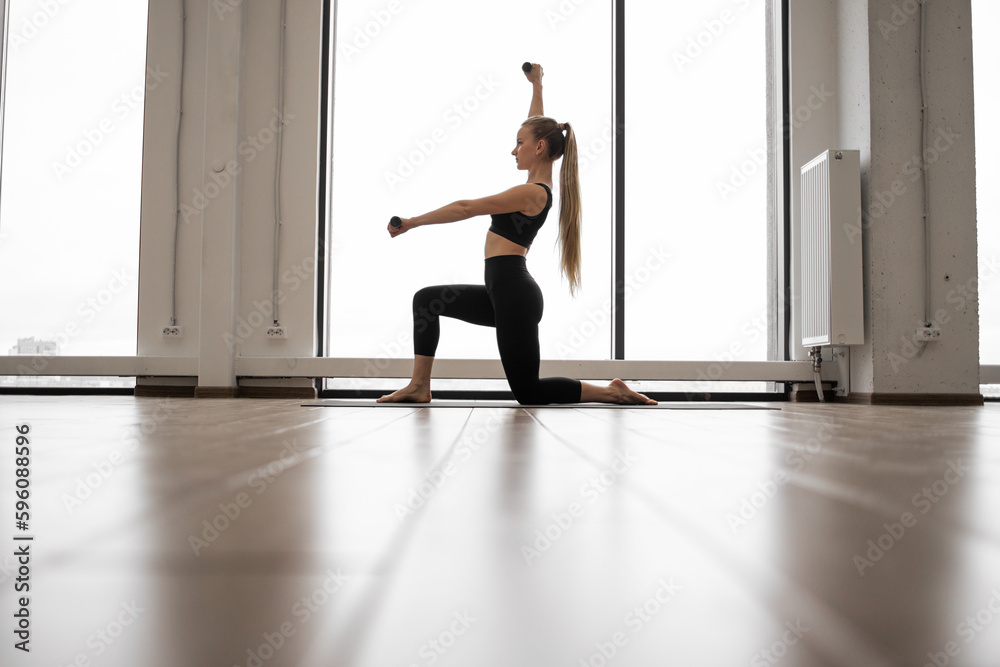 Strong caucasian woman standing with one knee on yoga mat and using dumbbells for arms exercises. Well-trained lady in black activewear having workout near panoramic windows with city view. Strong caucasian woman standing with one knee on yoga mat and using dumbbells for arms exercises. Well-trained lady in black activewear having workout near panoramic windows with city view.